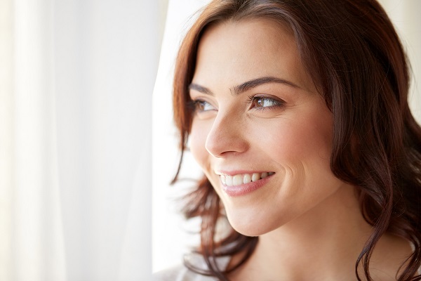 people and leisure concept - happy young woman looking through window at home