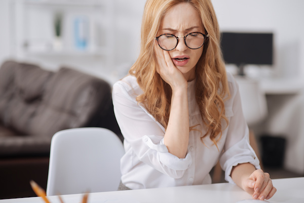 Need to see dentist. Puzzled upset young worker sitting at the workplace while touching her cheek and suffering from aching teeth