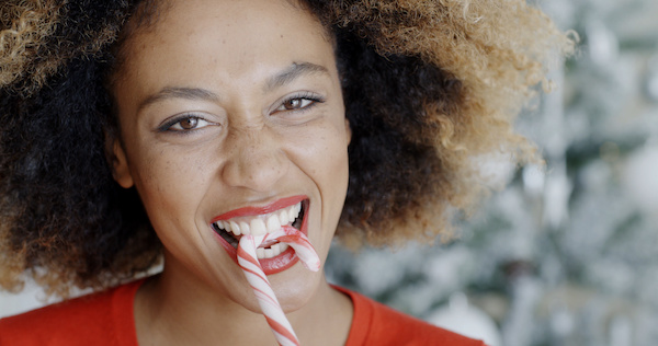Fun young woman biting a traditional red and white striped Christmas candy cane as she looks at the camera with a mischievous smile