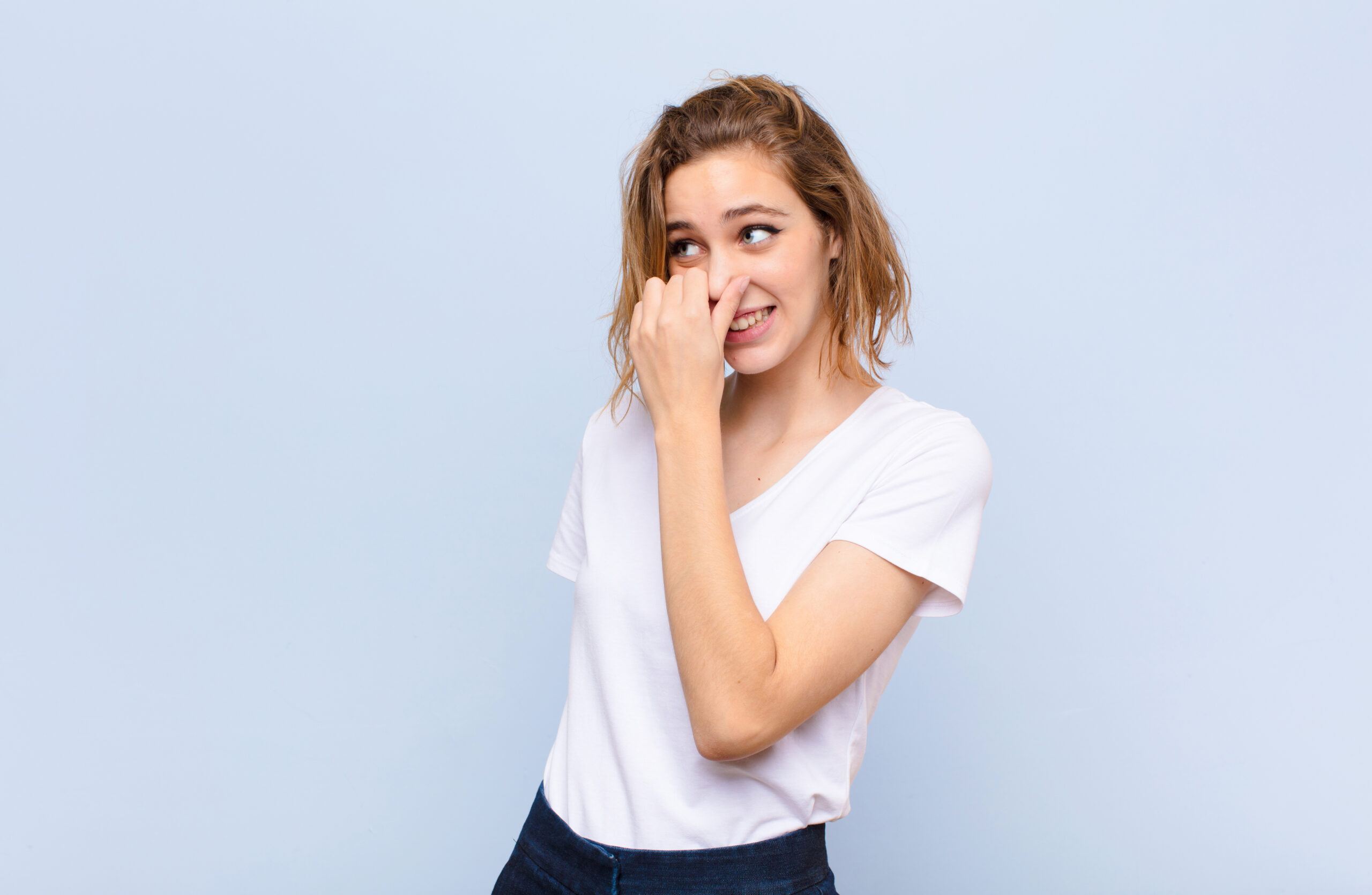 young blonde woman feeling disgusted, holding nose to avoid smelling a foul and unpleasant stench against flat color wall