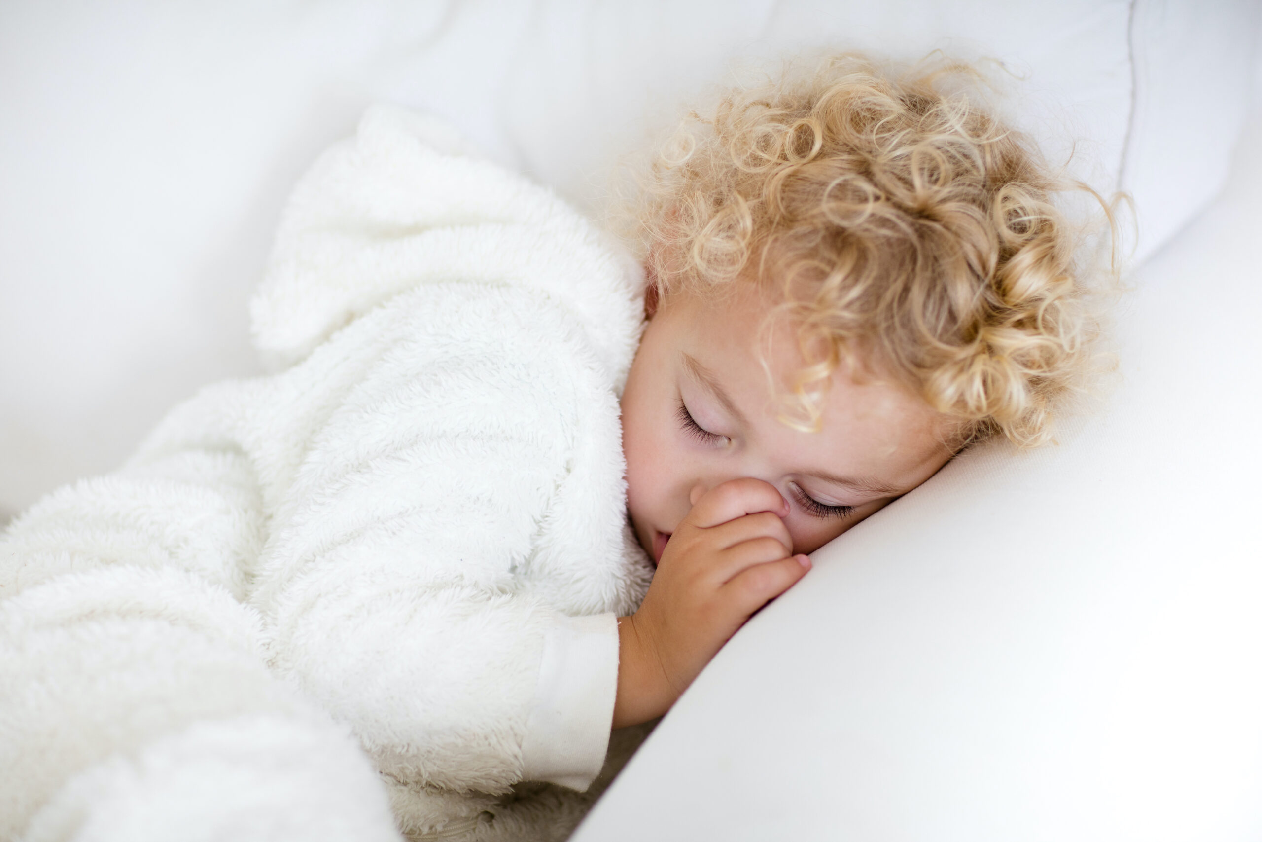 thumb sucking Cute blond curly little boy sleeping on white couch. Tired toddler taking a nap wearing warm white pajamas sucking his thumb.