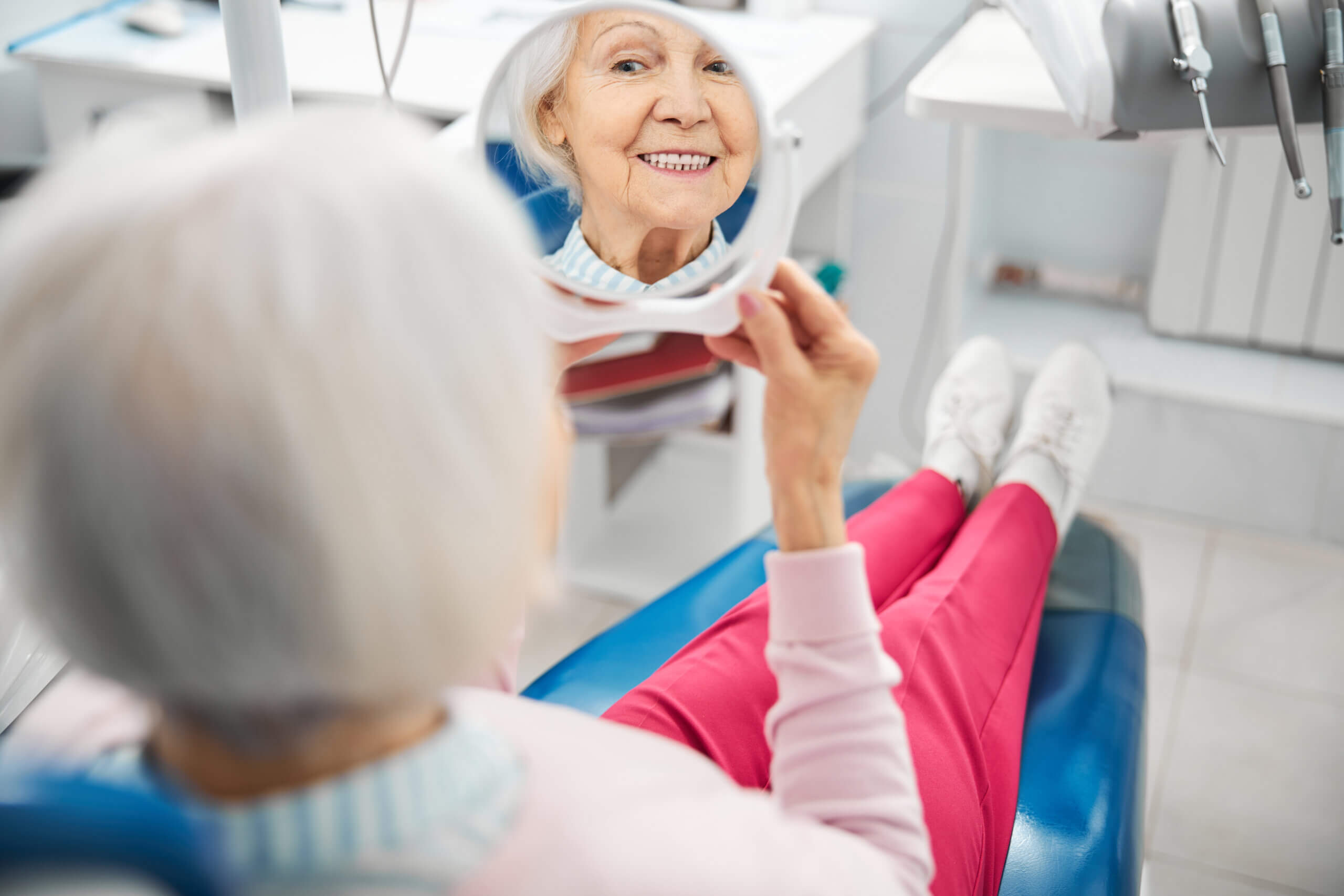 Pensioner looking at her reflection in a small mirror while showing teeth and lying in a dentist chair